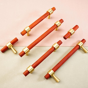 May include: A collection of wooden cabinet pulls with a reddish-brown finish and gold-toned metal accents. The pulls have a cylindrical design with a central band and a T-shaped handle. The hardware is arranged on a two-tone pink and beige background.