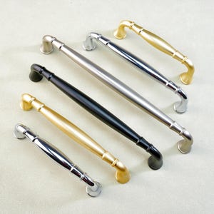 May include: An assortment of cabinet pulls in various finishes, including gold, silver, and black. The hardware features a curved handle design with rounded end mounts. The pulls are arranged diagonally on a light-colored surface.