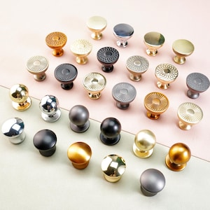 May include: A collection of cabinet knobs in a range of metallic finishes, such as gold, silver, and black. The knobs display diverse designs, from smooth spheres to textured patterns, arranged on a pink and green surface.