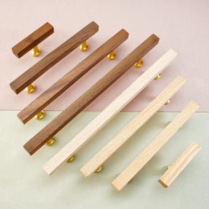 May include: A collection of wooden cabinet pulls and knobs in various shades of brown and natural wood. Each pull is rectangular with a brass-colored base. The pulls are arranged diagonally against a two-tone pink and green background.