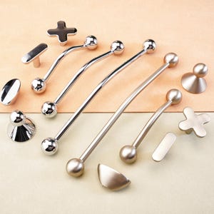 Könnte beinhalten: Eine Sammlung von silberfarbenen Schrankbeschlägen, darunter Griffe, Knöpfe und Züge. Das Sortiment umfasst verschiedene Formen, wie gebogene Griffe mit Kugelenden, kreuzförmige Knöpfe und ovale Züge. Die Hardware ist auf einer zweifarbigen beige-cremefarbenen Oberfläche angeordnet.