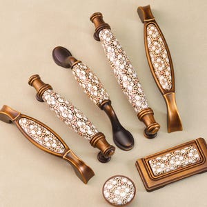 Op de afbeelding: Een verzameling decoratieve kastbeslagen, waaronder handgrepen en een knop. De hardware heeft een bronzen afwerking met witte bloempatroon keramische inzetstukken. De handgrepen variëren in vorm en grootte.