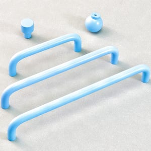 Pode incluir: Um conjunto de ferragens para armários em azul claro, incluindo três puxadores de barra curvados, um pequeno botão e um puxador esférico. Os itens estão dispostos em uma superfície cinza claro, mostrando um design minimalista.