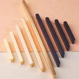 Op de afbeelding: Assortiment van gouden en zwarte kastbeslaggrepen in verschillende maten. De grepen zijn rechthoekig van vorm met afgeronde randen.