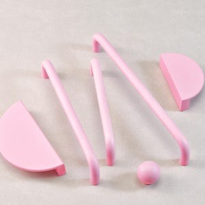 May include: A collection of pink cabinet hardware, including two semi-circular pulls, three curved bar pulls, and a small spherical knob. The hardware is made of a solid, matte pink material and is arranged on a light gray surface.