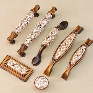 Op de afbeelding: Een verzameling decoratieve kastbeslagen, waaronder handgrepen en een knop. De hardware heeft een bruine afwerking met witte en bruine bloempatroon keramische inzetstukken. De handgrepen variëren in grootte en vorm.