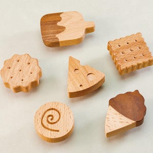 May include: A collection of six wooden food-shaped toys, including a cookie, cheese wedge, ice cream cone, and a biscuit. The toys are made of light-colored wood with some pieces featuring darker wood accents.