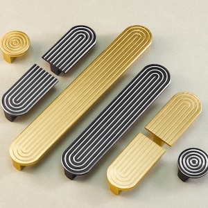 May include: An assortment of gold and black cabinet hardware, including knobs and pulls. The hardware features a grooved design with a curved, arched shape. The knobs have a circular design with concentric grooves.