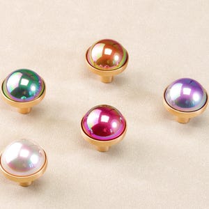 Peut inclure: Cinq boutons de meuble irisés avec une finition nacrée. Chaque bouton présente un dessus rond et coloré, semblable à du verre, et une base dorée. Les couleurs incluent le vert, le rouge, le violet et le blanc. Ces boutons décoratifs sont parfaits pour les meubles.