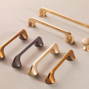 May include: An assortment of cabinet hardware in various metallic finishes. The collection includes several bar pulls and a round knob. Colors range from gold to bronze and dark gray. The hardware is arranged on a neutral surface.
