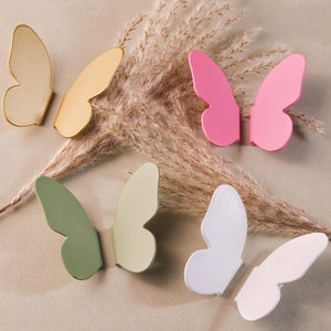 Może przedstawiać: Pięć dekoracyjnych haczyków ściennych w kształcie motyla w różnych kolorach, w tym złoty, zielony, biały i różowy. Haczyki są wykonane z metalu i mają prosty, nowoczesny design.