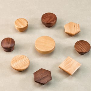 Może przedstawiać: Kolekcja drewnianych gałek do szafek w r&oacute;żnych kształtach i odcieniach drewna. Gałki obejmują okrągłe, sześciokątne i kwadratowe wzory, prezentujące naturalne wzory słoj&oacute;w drewna. Układ podkreśla r&oacute;żnorodność dostępnych styl&oacute;w.