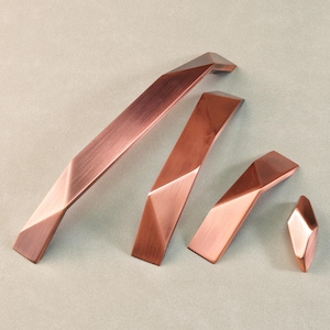 May include: A set of copper-toned cabinet handles and a knob. The handles vary in size, with a long bar and two shorter ones. They feature a geometric, faceted design. The knob is a small, faceted shape.