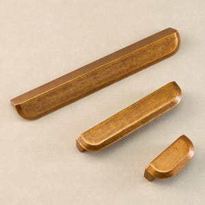 Op de afbeelding: Drie bronzen kastgrepen in verschillende maten. De grepen hebben een rechthoekige vorm met afgeronde uiteinden en een licht getextureerd oppervlak. Ze zijn diagonaal geplaatst op een neutrale achtergrond.