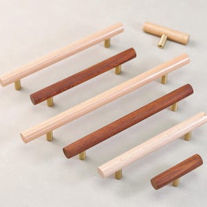 Könnte beinhalten: Holzschrankgriffe in verschiedenen Braun- und Naturholztönen. Die zylindrischen Griffe haben Messingfarbene Befestigungen. Die Kollektion umfasst lange Stangengriffe und einen kürzeren, T-förmigen Griff.