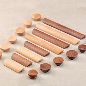 May include: A collection of wooden cabinet hardware, including knobs and pulls. The knobs are round, hexagonal, and mushroom-shaped, in various shades of light and dark brown. The pulls are rectangular with rounded edges.