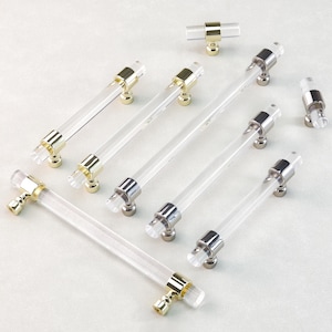 May include: Clear acrylic cabinet pulls with gold and silver-toned hardware. The pulls are cylindrical with a T-bar shape. The hardware includes a round base and a decorative accent. The hardware is available in gold and silver finishes.
