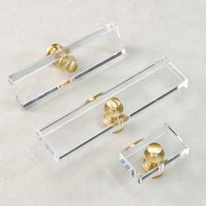 May include: Three clear acrylic cabinet pulls with brass hardware. The pulls are rectangular and vary in size, with a small square pull and two longer rectangular pulls. The brass hardware is circular and attached to the center of each pull.