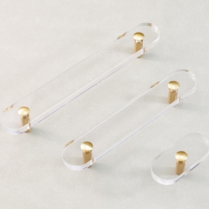 May include: Clear acrylic cabinet pulls with brass-colored hardware. The pulls are oval-shaped with rounded ends and are available in three different sizes. The hardware is a cylindrical shape. The pulls are designed for use on cabinets and drawers.