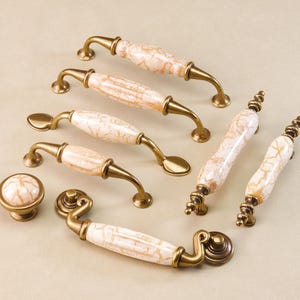 Peut inclure: Ensemble de quincaillerie d'armoire, comprenant des poignées et un bouton. Les poignées ont un centre en céramique crème marbrée avec des accents métalliques couleur laiton. Le bouton est rond avec un dessus en céramique assorti. Quincaillerie pour meubles.