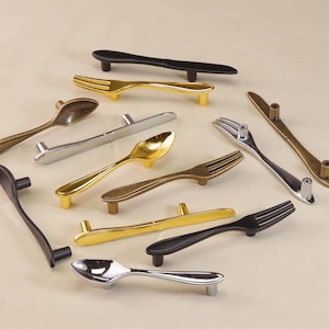 May include: A selection of decorative cabinet handles shaped like cutlery. The handles are in various metallic finishes, including gold, silver, and black. The assortment includes spoon, fork, and knife designs, adding a unique touch to furniture.