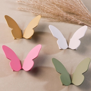 以下が含まれることがあります： 金、白、ピンク、緑の蝶の形をした4つのキャビネットノブ。 各ノブは、家具に気まぐれなタッチを加えるのに最適な、一対の様式化された翼を備えています。 ノブは金属製です。