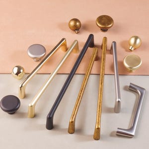 Könnte beinhalten: Sortiment an Schrankbeschlägen in verschiedenen Ausführungen, darunter Gold, Silber und Schwarz. Die Beschläge umfassen Knöpfe und Griffe in runden, quadratischen und rechteckigen Formen.