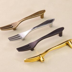 May include: Four cabinet pulls designed as forks, in antique brass, silver, dark bronze, and gold finishes. Each pull has a fork-shaped handle and a small post for mounting. The pulls are designed for use on cabinets and drawers.