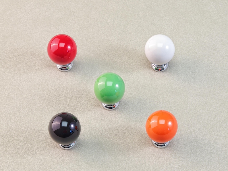 Peut inclure: Cinq boutons de meuble color&eacute;s en rouge, blanc, vert, noir et orange. Chaque bouton est une sph&egrave;re brillante mont&eacute;e sur une base argent&eacute;e. Les boutons sont dispos&eacute;s sur une surface neutre de couleur claire.