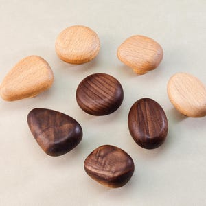 May include: A collection of wooden cabinet knobs in various shapes and shades. Some are light-colored, while others are a rich, dark brown. The knobs are arranged on a neutral background, showcasing their natural wood grain and unique forms.