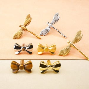 May include: A selection of decorative metal drawer pulls and cabinet knobs. The collection features dragonfly and bow-shaped designs in gold, silver, and bronze finishes. The hardware is displayed on a peach-coloured surface.