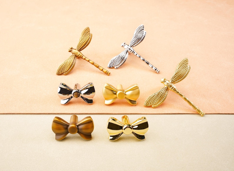 May include: A selection of decorative metal drawer pulls and cabinet knobs. The collection features dragonfly and bow-shaped designs in gold, silver, and bronze finishes. The hardware is displayed on a peach-coloured surface.