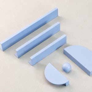 Può includere: Una collezione di ferramenta per mobili azzurri, tra cui maniglie rettangolari e semicircolari e una piccola sfera. L'hardware è disposto su una superficie neutra, mostrando un design moderno e minimalista.