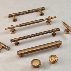 Könnte beinhalten: Eine Auswahl an antiken Messing-Schrankbeschlägen, darunter Stangengriffe, T-förmige Griffe und runde Knöpfe. Die Beschläge weisen strukturierte Oberflächen und eine warme, gealterte Oberfläche auf, geeignet für Küchen- oder Möbel-Upgrades. Maße in Zentimetern.