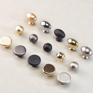 May include: A collection of cabinet knobs in diverse metallic finishes, including gold, silver, and black. The knobs are in various shapes, such as round, oval, and spherical, and are arranged on a light-coloured surface.