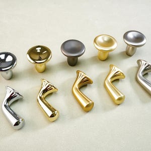May include: An assortment of cabinet knobs and pulls in various metallic finishes. The knobs are round with a mushroom-shaped base, while the pulls have a curved, horn-like design. Colors include silver, gold, and a dark gray.