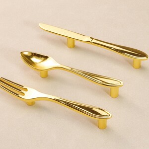 May include: A set of gold-coloured cabinet handles, each shaped like a piece of cutlery: a knife, a spoon, and a fork. The handles are mounted on short, cylindrical posts. The shiny, metallic material suggests brass or a similar alloy.