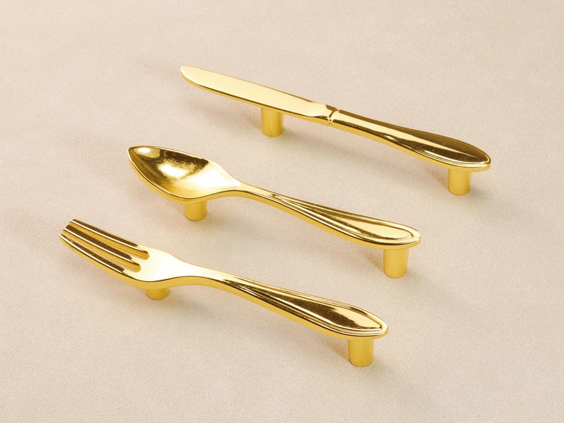 May include: A set of gold-coloured cabinet handles, each shaped like a piece of cutlery: a knife, a spoon, and a fork. The handles are mounted on short, cylindrical posts. The shiny, metallic material suggests brass or a similar alloy.