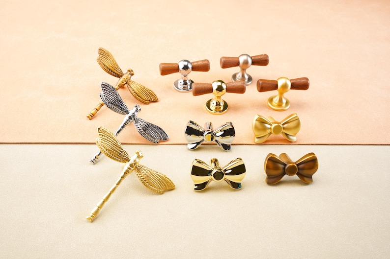 May include: A selection of decorative cabinet hardware, including dragonfly, bow tie, and bar-style knobs. The hardware is in various metallic finishes, such as gold, silver, and bronze. The knobs are designed for furniture and home decor.