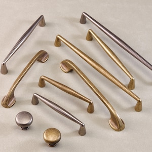 Op de afbeelding: Een assortiment bronzen en zilverkleurige kastbeslagen, waaronder handgrepen en knoppen. De handgrepen variëren in grootte en vorm, sommige hebben een gebogen ontwerp. De knoppen zijn rond en hebben een bijpassende afwerking.