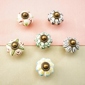 May include: Six decorative ceramic cabinet knobs with floral designs. The knobs feature a variety of colours, including white, green, black, and blue, with gold-coloured metal accents. The knobs are arranged on a pink and beige background.