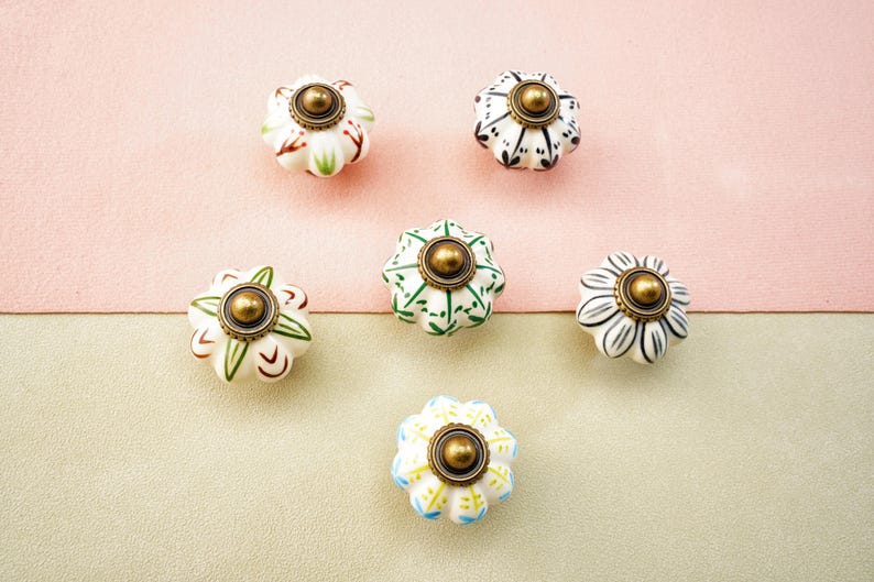 May include: Six decorative ceramic cabinet knobs with floral designs. The knobs feature a variety of colours, including white, green, black, and blue, with gold-coloured metal accents. The knobs are arranged on a pink and beige background.