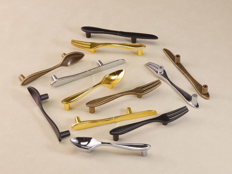 May include: A selection of decorative cabinet handles shaped like cutlery. The handles are in various metallic finishes, including gold, silver, and black. The assortment includes spoon, fork, and knife designs, adding a unique touch to furniture.