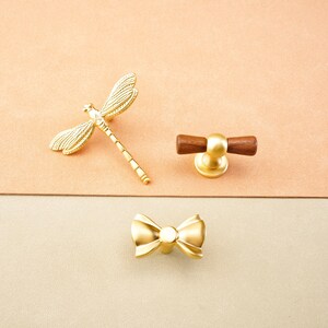 May include: Three decorative cabinet knobs in a gold finish. One is a dragonfly, another a bow, and the third has a wooden handle. The knobs are displayed on a two-tone background.
