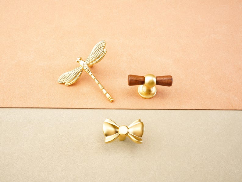 May include: Three decorative cabinet knobs in a gold finish. One is a dragonfly, another a bow, and the third has a wooden handle. The knobs are displayed on a two-tone background.