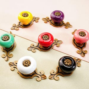 May include: A collection of seven colorful cabinet knobs. Each knob features a round, glossy ceramic top in yellow, purple, red, pink, green, white, and black. The knobs are mounted on ornate, antique bronze-colored metal backplates.