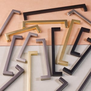 May include: Assortment of cabinet hardware pulls in various finishes, including black, gold, and nickel. The pulls are all rectangular in shape with a slight curve at the top and bottom.