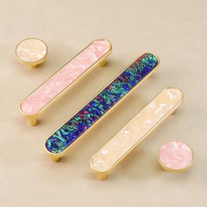May include: Decorative cabinet hardware set featuring a gold finish. The set includes two round knobs and three rectangular pulls with colorful, marbled inserts in pink, blue, and cream. The hardware is designed for furniture and home decor.