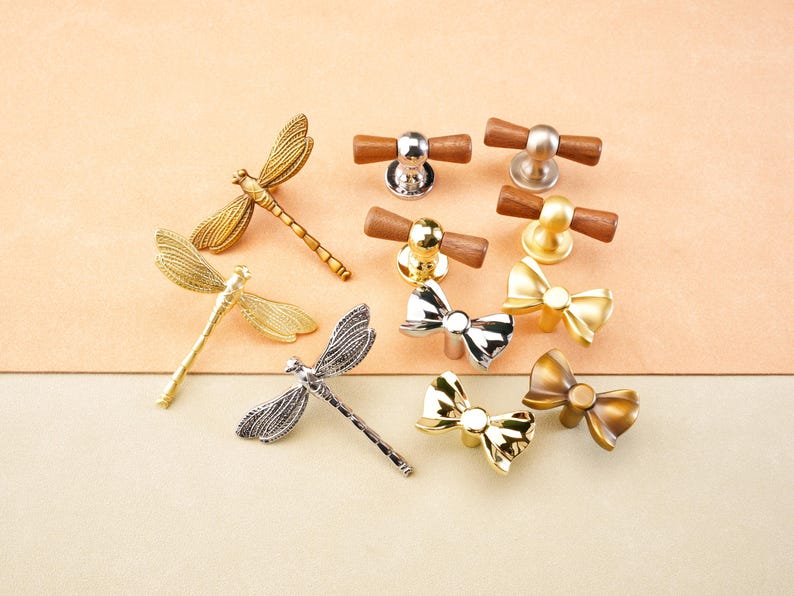 May include: A selection of cabinet knobs and pulls in various designs. The collection includes dragonfly, bow, and wooden bar styles. Finishes include gold, silver, and bronze tones. The knobs are set against a neutral background.