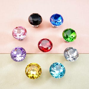 Op de afbeelding: Een verzameling van negen kleurrijke kristallen kastknoppen, gerangschikt in een cirkel. De knoppen zijn in verschillende kleuren, waaronder zwart, blauw, roze, groen, rood, paars, zilver, geel en turquoise. Elke knop heeft een facetontwerp.